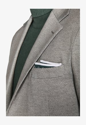 Grijze herringbone blazer met notch lapels, gecombineerd met een donkergroene geribde coltrui. Zakdoek in wit en groen zichtbaar in de borstzak.