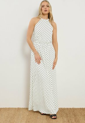 Kvinde stående iført en hvid ærmeløs halterneck maxi-kjole med sorte polkaprikker og sorte sandaler med åben tå på et vævet tæppe.