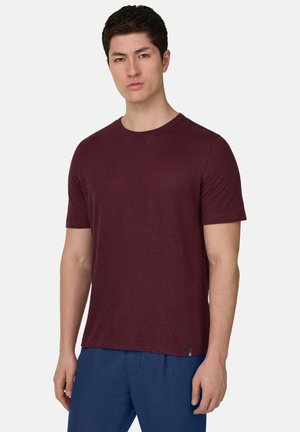 Junger Mann mit kurzen dunklen Haaren, der ein schlichtes bordeauxrotes T-Shirt mit kurzen Ärmeln und blaue Hose trägt, vor einem weißen Hintergrund stehend.