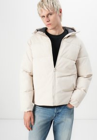 Chaqueta acolchada beige con capucha, que presenta un cierre de cremallera, paneles cosidos y puños elásticos, combinada con jeans de mezclilla azul.