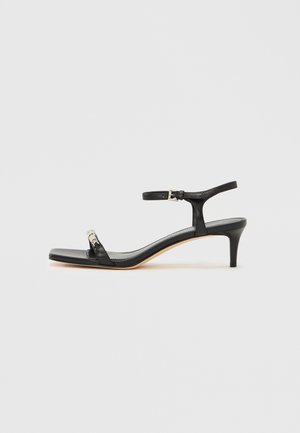 Sandale femme en cuir noir avec petit talon kitten, fine bride à la cheville avec boucle et bride unique sur les orteils avec petit détail métallique.