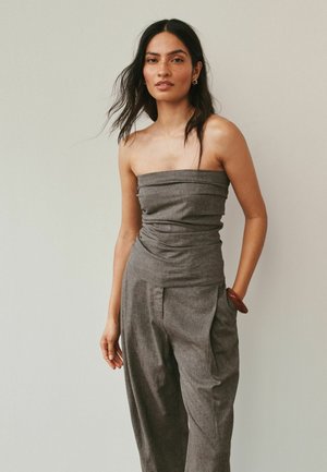 Kobieta o długich ciemnych włosach, ubrana w szary kombinezon bez ramiączek i brązową bransoletkę, stoi na jednolitym jasnobeżowym tle.