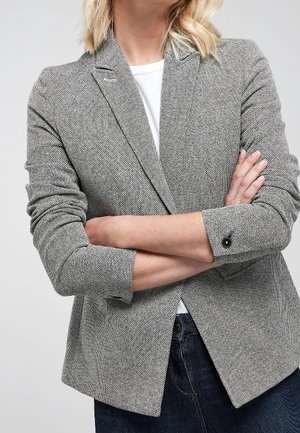 Frau trägt einen grauen Fischgrät-Blazer mit hochgekrempelten Ärmeln, weißem Hemd darunter, verschränkten Armen und dunkelblauen Jeans.