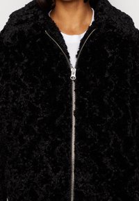 Svart fuskpälsjacka med en mjuk, texturerad yta, med en silverfärgad dragkedja och en avslappnad passform. Inga synliga mönster eller detaljer.