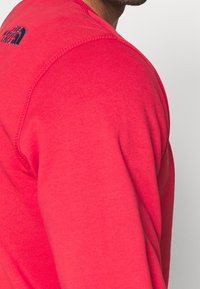 Czerwony sweter z bawełny, z dekoltem w serek, widocznymi szwami i czarnym logo na górnej części pleców. Miękka faktura.
