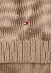 Maglione beige lavorato a maglia con texture a coste, modelli di maglia orizzontali e verticali a contrasto, e un piccolo logo rosso, bianco e blu.