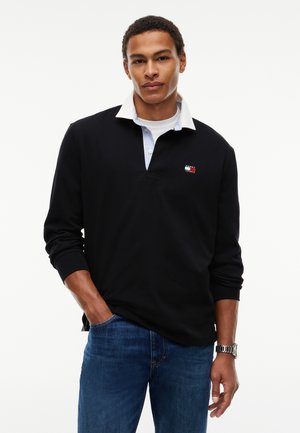Ung mand med kort krøllet hår iført en sort langærmet skjorte med en hvid krave og blå jeans, står med den ene hånd i lommen.