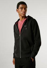 Μαύρο φούτερ με φερμουάρ και κορδόνια, μπροστινές τσέπες και ραμμένα μανίκια. Φοριέται πάνω από ένα κόκκινο μπλουζάκι, που διαθέτει λευκή κεντημένη εκτύπωση στο στήθος.