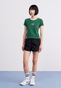 Žalias Nike marškinėliai su balta apykakle, juodi šortai ir balti sportbačiai. Modelis vilki juodai baltas dryžuotas kojines. Paprastas, sportinis dizainas.