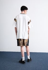 Camisa branca de manga curta com mangas em padrão de camuflagem, combinada com calções camuflados castanhos. Ténis pretos e meias brancas até ao tornozelo. Fundo simples.