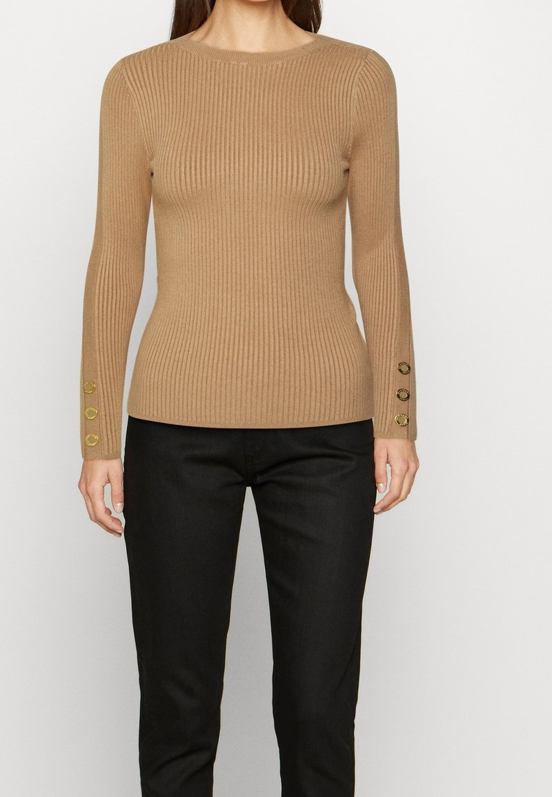 Femme portant un pull camel près du corps en côtes avec des boutons dorés sur les manches et un pantalon noir sur un fond uni.