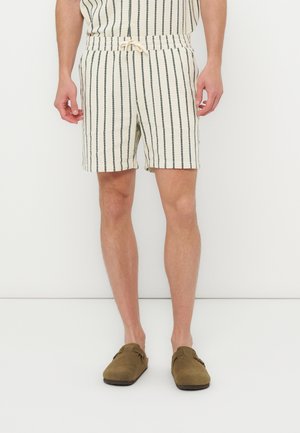 Homme portant un short beige à rayures noires verticales et des chaussures marron sans lacets, debout devant un fond blanc uni.