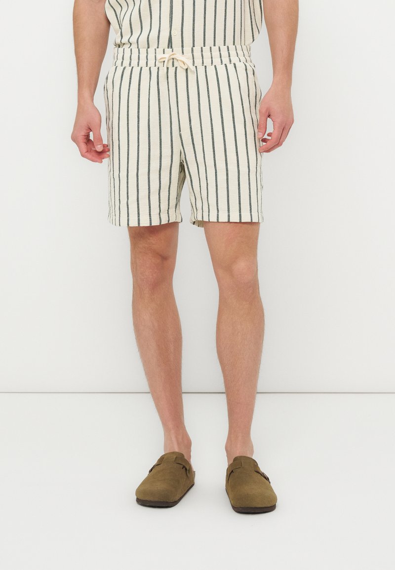 Homme portant un short beige à rayures noires verticales et des chaussures marron sans lacets, debout devant un fond blanc uni.