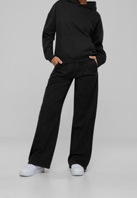 Persona con pantalones negros holgados y una sudadera negra a juego, con las manos en los bolsillos, combinada con zapatillas blancas, sobre un fondo gris.