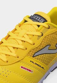 Κίτρινο αθλητικό παπούτσι με επάνω μέρος από δίχτυ, κίτρινες κορδόνια, μαύρο και γκρι λογότυπο, και την επιγραφή "Mundial" στο πλάι κοντά στην περιοχή των δαχτύλων.