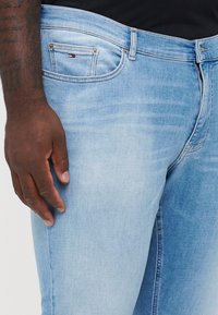 Light blue denim jeans with a slim fit, featuring front pockets, subtle fading, and branded accents near the pocket. Smooth texture.