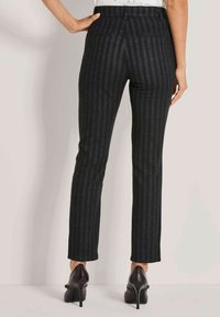 Vrouw in een strakke zwarte pinstripe broek en zwarte hoge hakken, staande met één hand op haar heup tegen een effen muur.