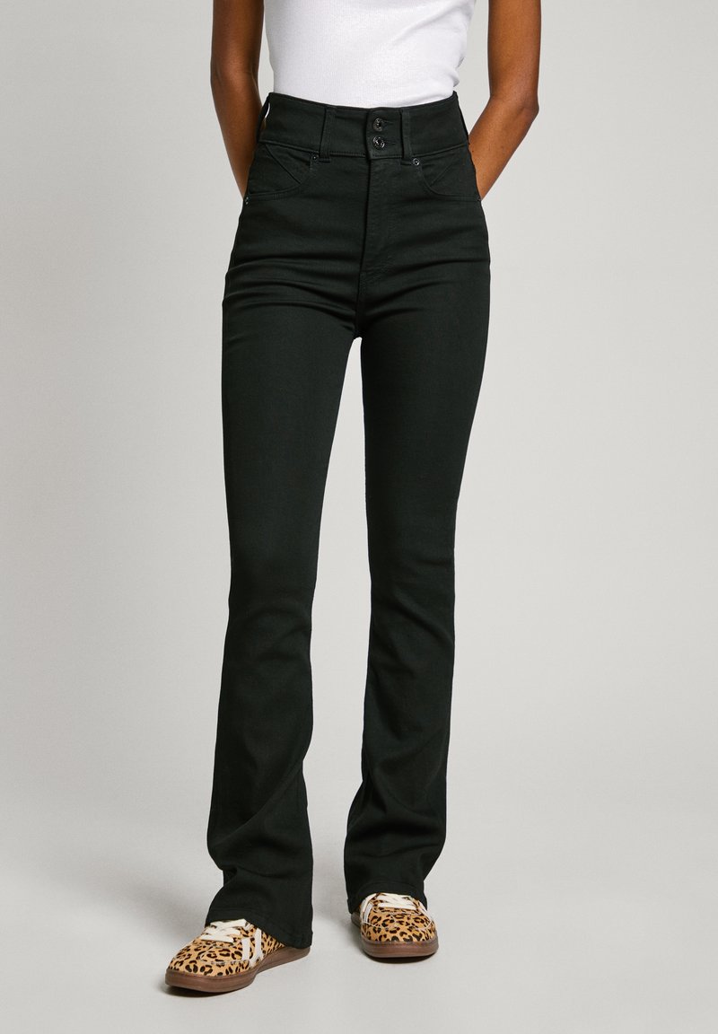 Personne portant un pantalon noir évasé taille haute, un haut blanc et des baskets à imprimé léopard, debout devant un fond uni.