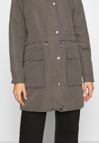 Kaki parka-jacka med medellång längd, utrustad med två framfickor, tryckknappsstängning och justerbara dragsnören i midjan.