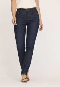 Jean en denim bleu foncé avec une coupe slim, présentant des surpiqûres dorées, une fermeture par bouton et des poches avant. Texture lisse et jambes droites.