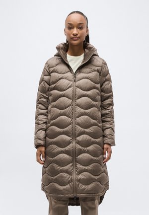 Žena stoji, nosi duguljasti smeđi prošiveni zimski kaput s visokim ovratnikom i prednjim patentnim zatvaračem, neutralna pozadina.