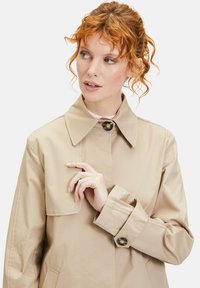 Beige Trenchcoat mit klassischem Kragen, großer Knopfverschluss und Manschettendetails. Glattes Material mit leichtem Glanz. Geeignet zum Schichten.