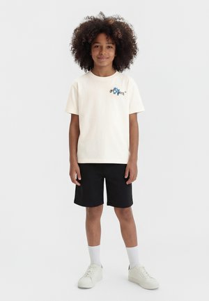 Παιδί με σγουρά μαλλιά στέκεται με το πρόσωπο προς τα εμπρός, φοράει ανοιχτόχρωμο t-shirt με σχέδιο, μαύρα σορτς, λευκές κάλτσες και λευκά αθλητικά παπούτσια, μπροστά από απλό φόντο.