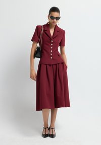 Donna che indossa un blazer corto buttoned bordeaux con maniche corte e una gonna midi a gamba larga coordinata, tacchi a punta neri, borsa nera, occhiali da sole a forma di gatto, orecchini dorati.
