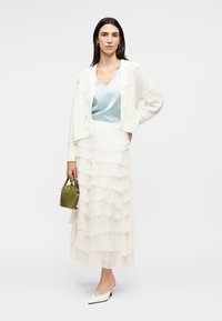 Γυναίκα που φορά λευκή φούστα με στρώσεις, ανοιχτό μπλε τοπ, λευκή ζακέτα, λευκές γόβες, κρατώντας μια μικρή τσάντα σε ελιά πράσινο χρώμα.