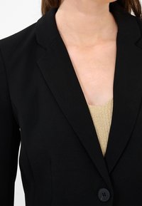 Svart kavaj med skarpa slag, enkel knapplåshållning och slät struktur, bärs över en guldstickad topp. Närbild av halsringningen.