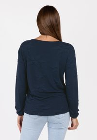 Top de manga larga en color navy con diseño floral texturizado, escote redondo y silueta ajustada. Combinado con unos jeans de color azul claro, mostrando un look casual.