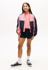 Μπουφάν αμάνικο σε ναυτικό μπλε και ροζ με ελαστικές μανσέτες, συνδυασμένο με ροζ φούτερ και ναυτικό σορτς. Άσπρα και ναυτικά sneakers ολοκληρώνουν την εμφάνιση.