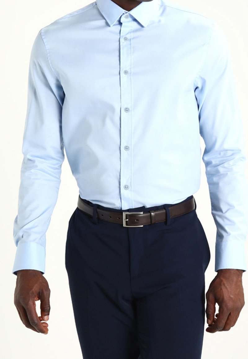 Homme portant une chemise habillée bleu clair boutonnée rentrée dans un pantalon bleu marine foncé avec une ceinture en cuir noire, debout devant un fond uni.
