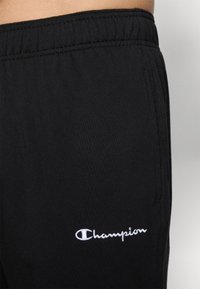 Schwarze Sporthose aus weichem Stoff, mit einem elastischen Bund und einem kleinen weißen Champion-Logo in der Nähe der Hüfte.