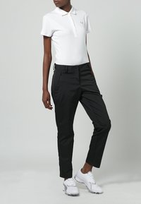 Vit polo med krage och logotyp, ihopparat med svarta avsmalnande byxor. Skorna är vita sneakers med grå detaljer.