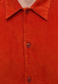 Gros plan d’une personne portant une chemise en velours côtelé rouge avec un col et trois boutons visibles à l’avant.