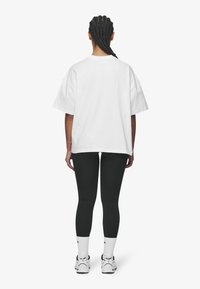 Bílé oversized tričko s kleslými rameny, spárované s černými legínami. Tenisky jsou bílé s černými akcenty a modelka má pletené vlasy.