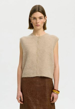 Donna con capelli corti castani che indossa un maglione beige senza maniche abbottonato, gonna in pelle marrone, orecchini a cerchio dorati e tre braccialetti spessi.