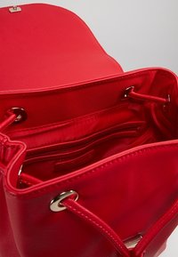 Bolso sintético rojo con una textura suave, cierre de cordón, bolsillos interiores y herrajes en tono plateado. Interior completamente forrado en tela roja.