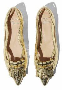 Złote metaliczne baleriny z zakończeniem w szpic, z wyciętymi bokami i akcentem kokardki z przodu. Teksturowana powierzchnia z widocznymi pęknięciami.