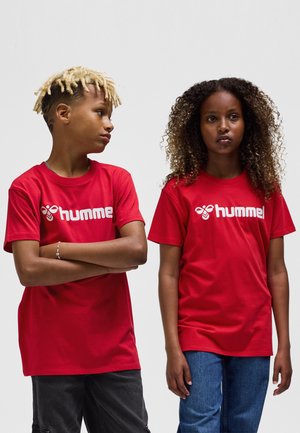 Röda bomulls-T-shirts med vitt "hummel"-logotyp, kortärmade och med avslappnad passform. En tröja har rund halsringning; synligt texturerat hår på modellerna.
