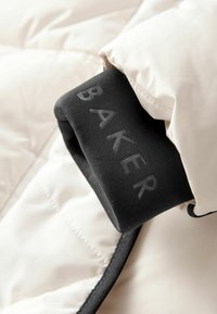 Helle puffer Jacke mit einem runden schwarzen Bündchen, auf dem das Wort "BAKER" in Grau steht. Sie hat eine gesteppten Struktur und schwarze Akzente.