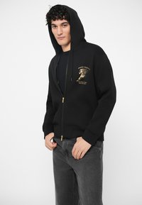 Jaunas vyras, vilkintis juoda užtraukiama džinsine striuke su gobtuvu, su auksiniu arkliu grafiku ir tekstu ant krūtinės, derinamas su tamsiai pilkais džinsais, pozuoja patalpoje.