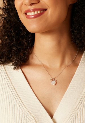 Zilveren ketting met een hartvormige pendant met een glanzende afwerking, hangend aan een delicate ketting, tegen een crèmekleurig top.