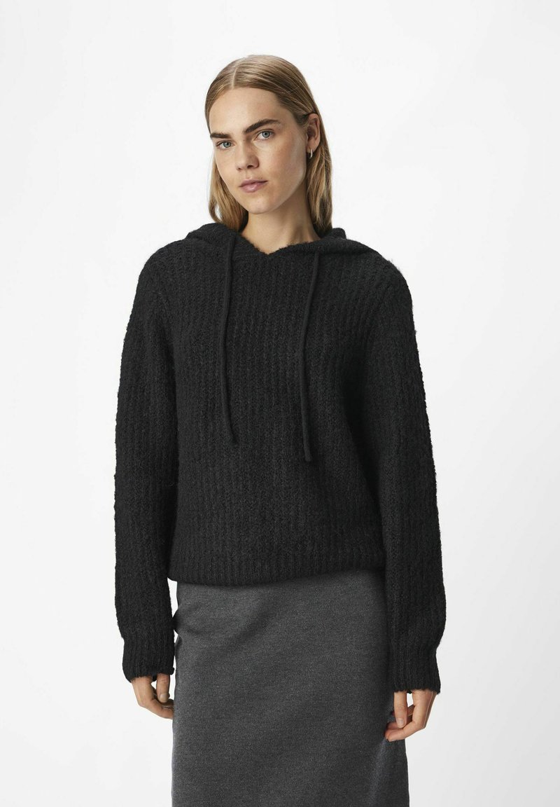 Zwarte hoodie met ribbeltextuur, lange mouwen en een touwtje. Gecombineerd met een grijze rok, op een neutrale achtergrond.