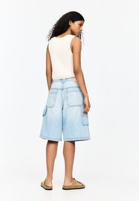 Helle blaue Denim-Cargo-Shorts mit Seitentaschen, kombiniert mit einem cremefarbenen, gerippten Tanktop. Das Modell wird von hinten mit welligem, dunklem Haar gezeigt.