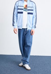 Ljust blå zip-up träningsjacka med mörkblå ränder, vit grafisk T-shirt under, lösa blåjeans och vita sneakers.