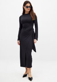 Black long-sleeve wrap dress in stretchy material, featuring a side tie detail and a fitted silhouette that falls to mid-calf.