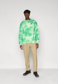 Φούτερ με γραβάτα-βαφή σε πράσινο χρώμα και ριμπ μανσέτες, μπεζ παντελόνι και μαύρα παπούτσια με λευκή σόλα. Casual σύνολο με κεντημένο λογότυπο.