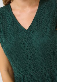 Grön klänning med V-ringning. Tyget har ett strukturerat geometriskt mönster och intrikata spetsdetaljer längs halsringningen.
